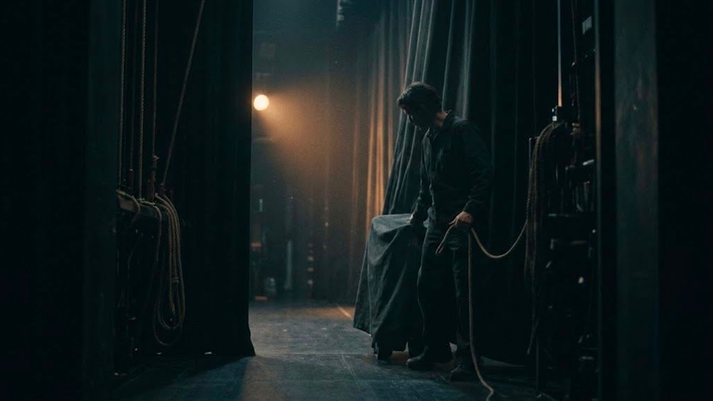 A dark, minimalist scene showing a glowing, arched doorway with a shadowy figure standing inside, partially reflected on a glossy floor, creating a mysterious and atmospheric mood.