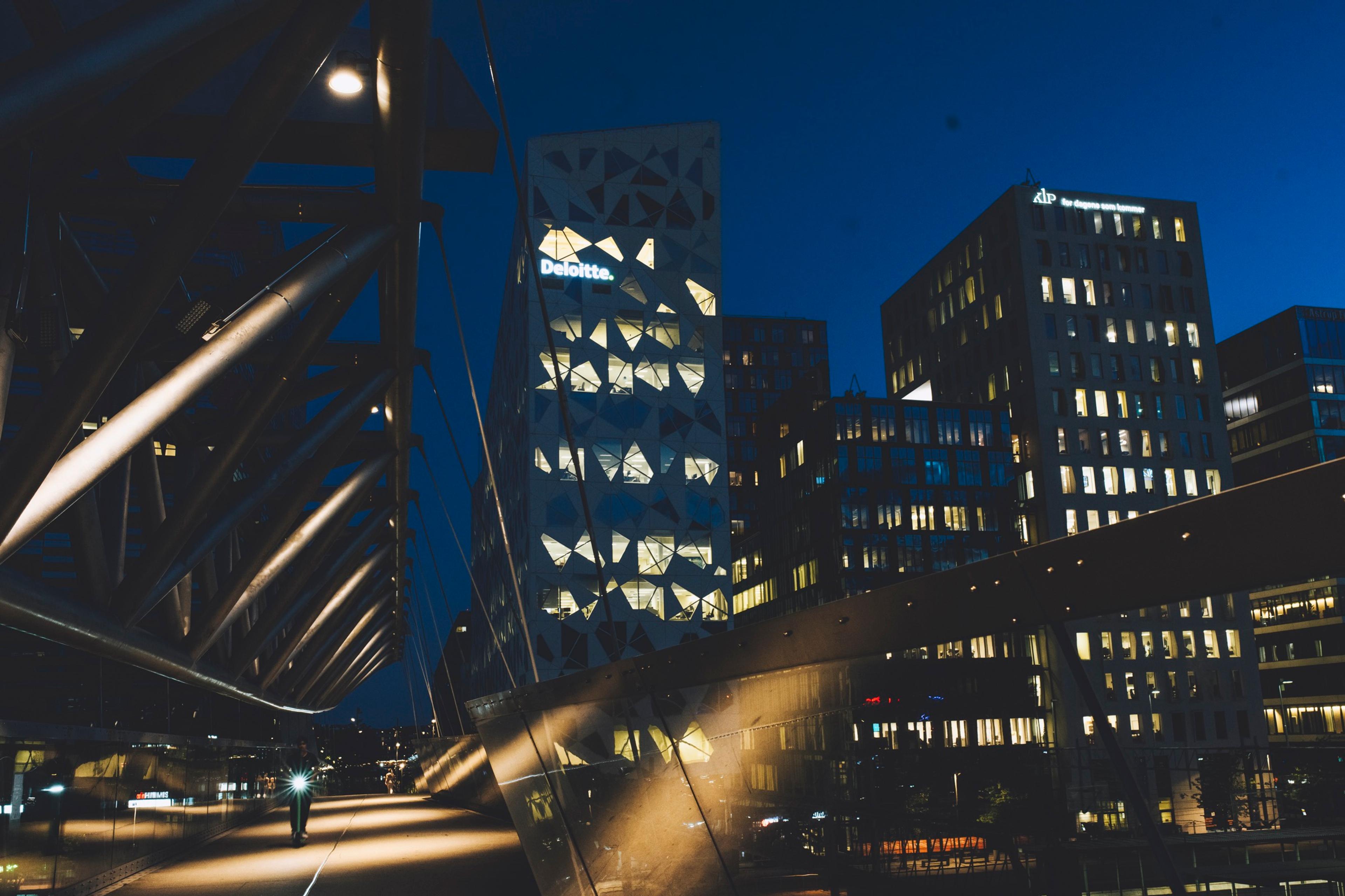 Det opplyste Deloitte-bygget i Bjørvika i Oslo om kvelden, med en gangbro i stål i forgrunnen.