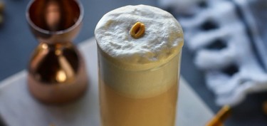 a cup of glass filled with a peanut colored drink with a white foam on top.