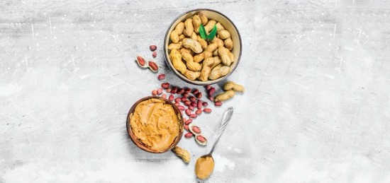 a bowl filled with unshelled peanuts next to a spoon with peanut butter.