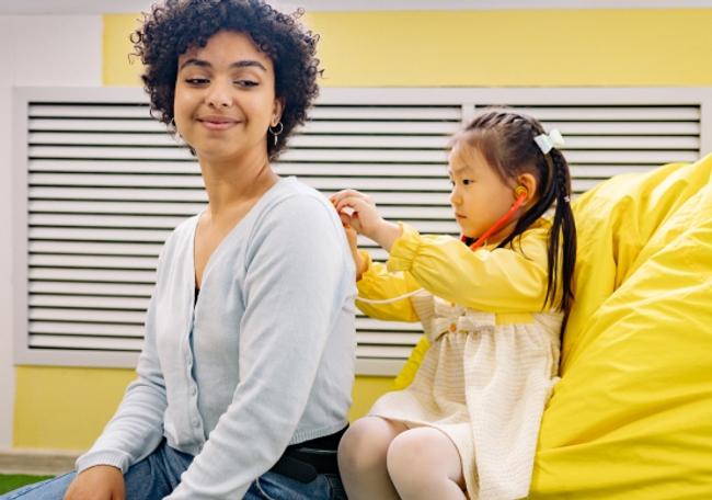 A woman is sitting on a yellow bean bag chair, facing back from a little girl, while the little girl using a stethoscope on the woman's back