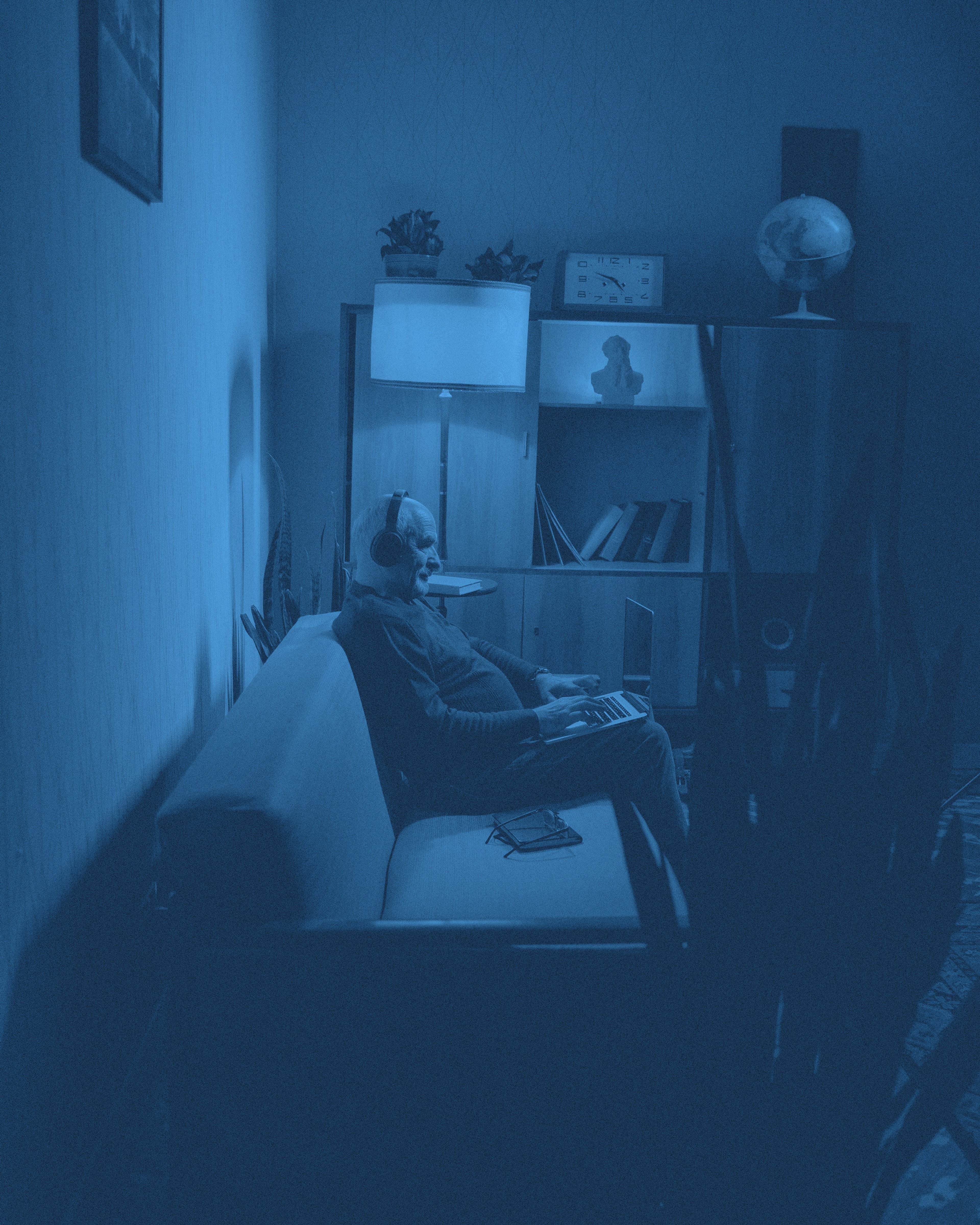 En äldre man sitter i en soffa, med hörlurar och dator