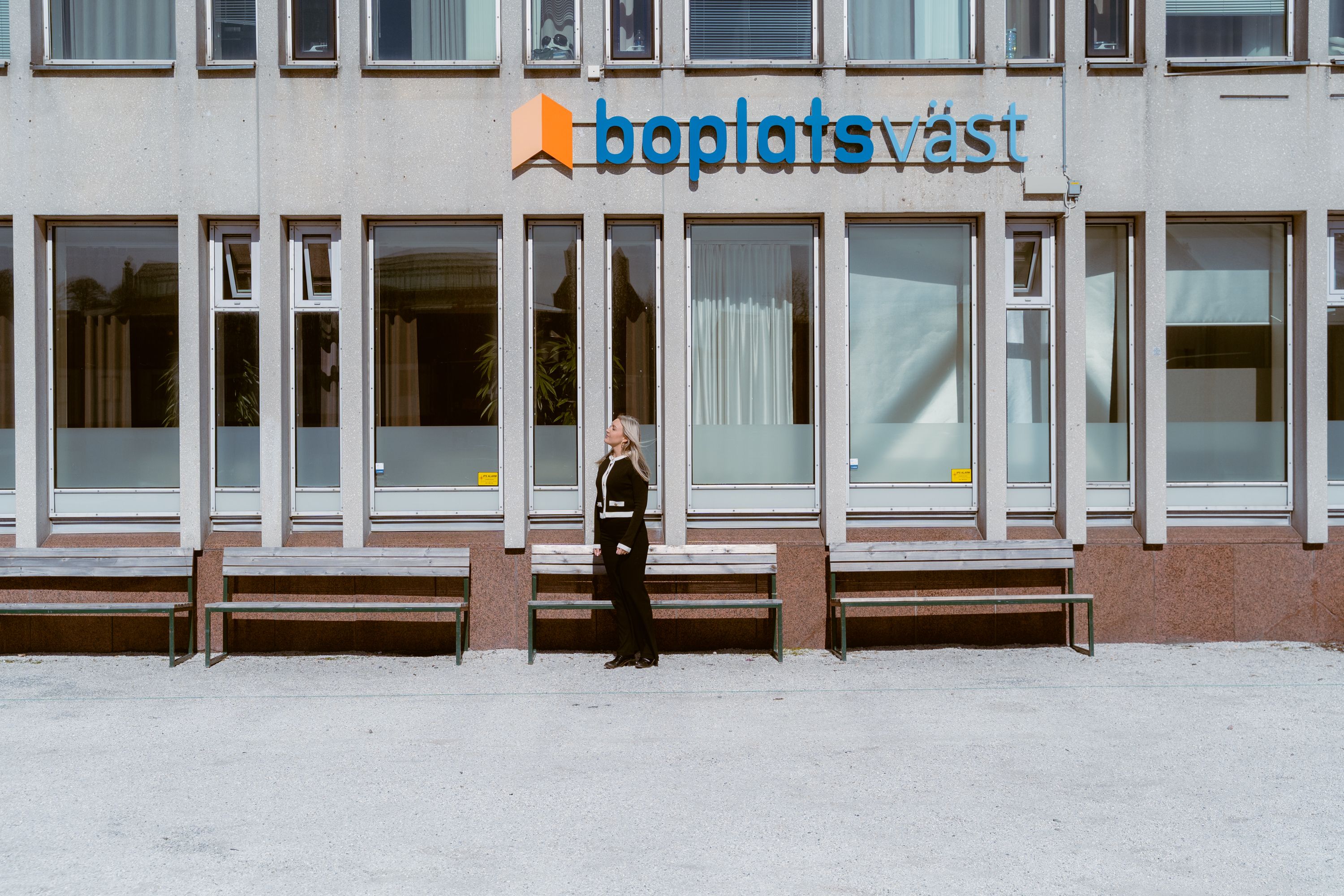 En människa står bredvid en husvägg, under en skylt där det står Boplats Väst