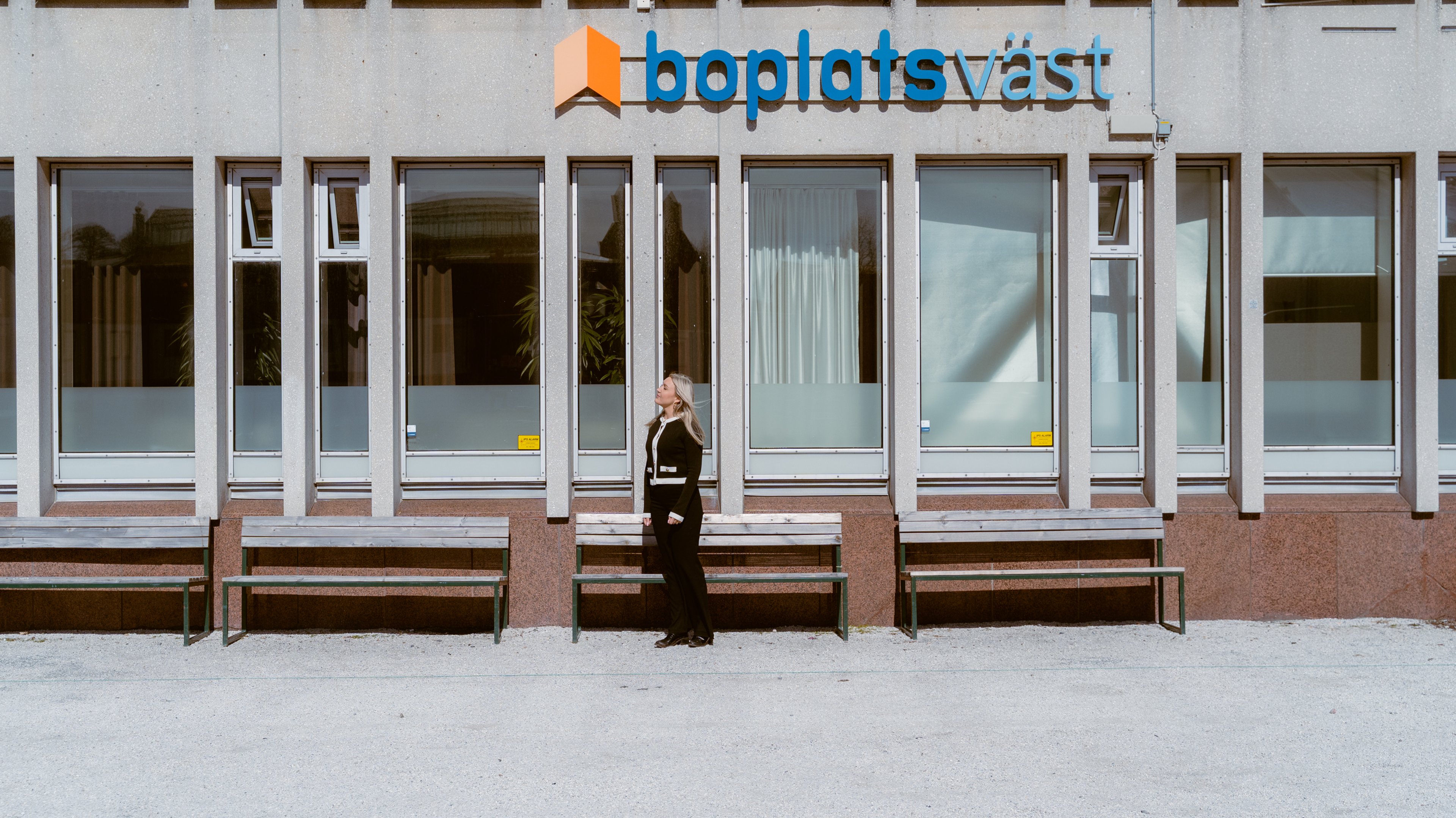 En människa står bredvid en husvägg, under en skylt där det står Boplats Väst