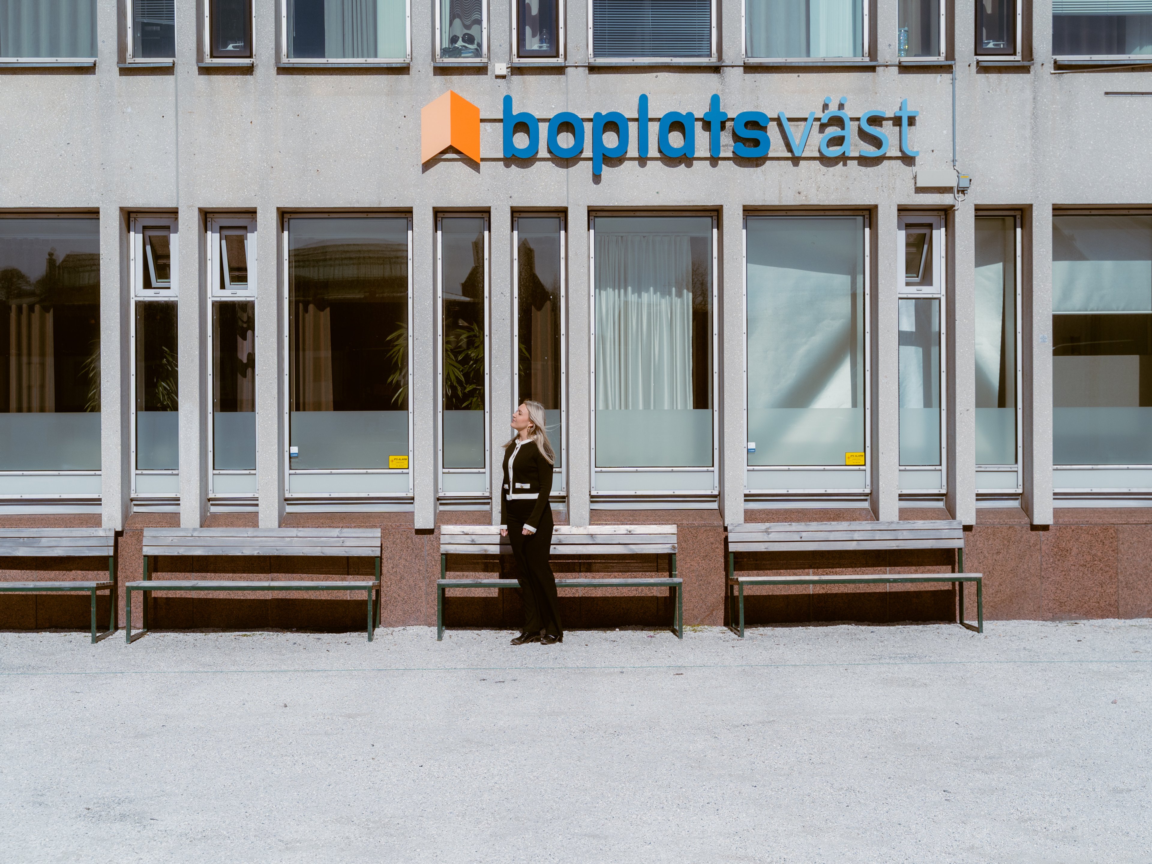 En människa står bredvid en husvägg, under en skylt där det står Boplats Väst