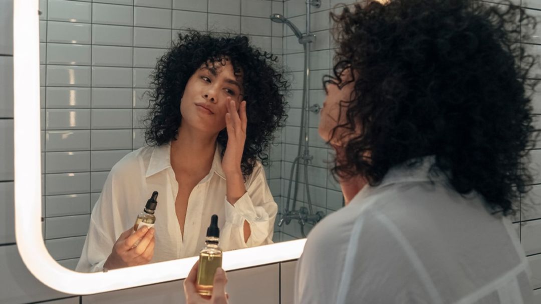 Young woman stood in bathroom setting looking in mirror and applying skincare to face