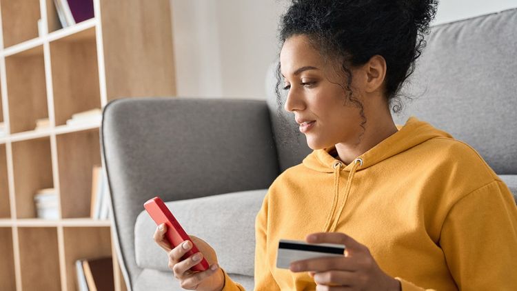Young adult female wearing casual clothing in a living room, using credit card and mobile phone.