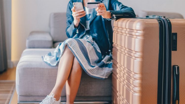 One adult woman sitting indoors with a suitcase and luggage, holding credit card and smart phone.