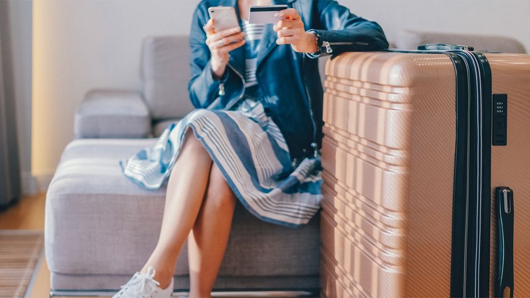 One adult woman sitting indoors with a suitcase and luggage, holding credit card and smart phone.