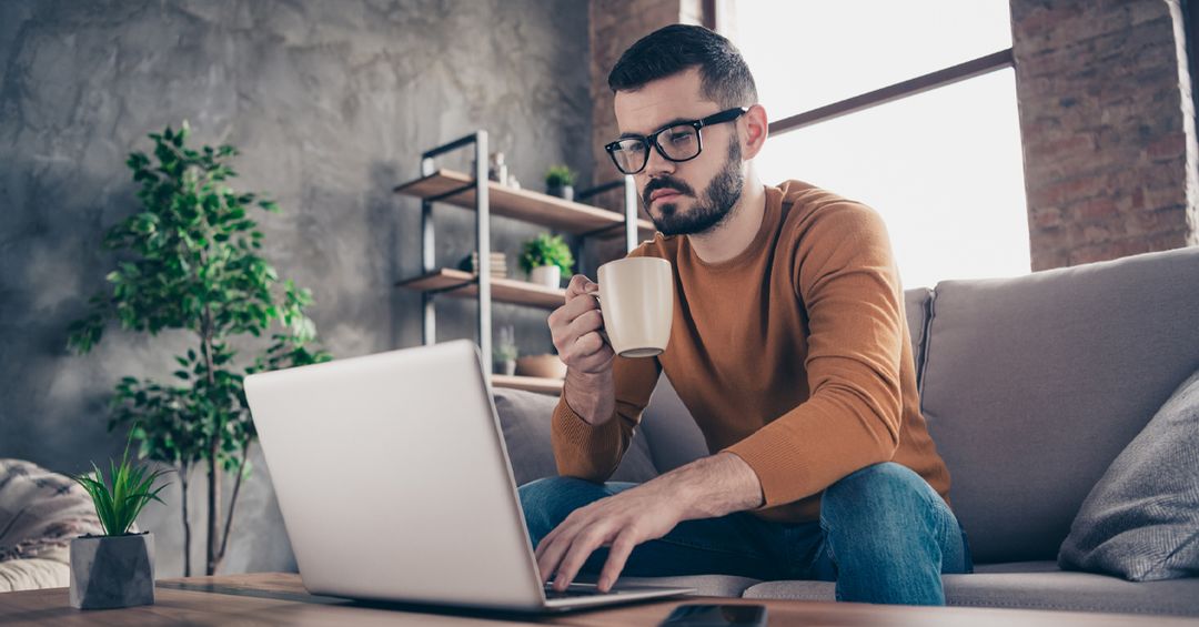 Man drinking coffee sitting on sofa using laptop in home setting