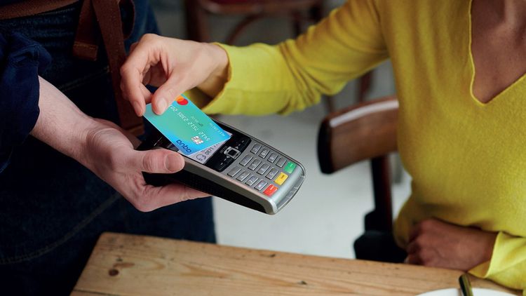 Close-up of adult woman using an Aqua credit card to pay at a contactless credit card reader in a café.
