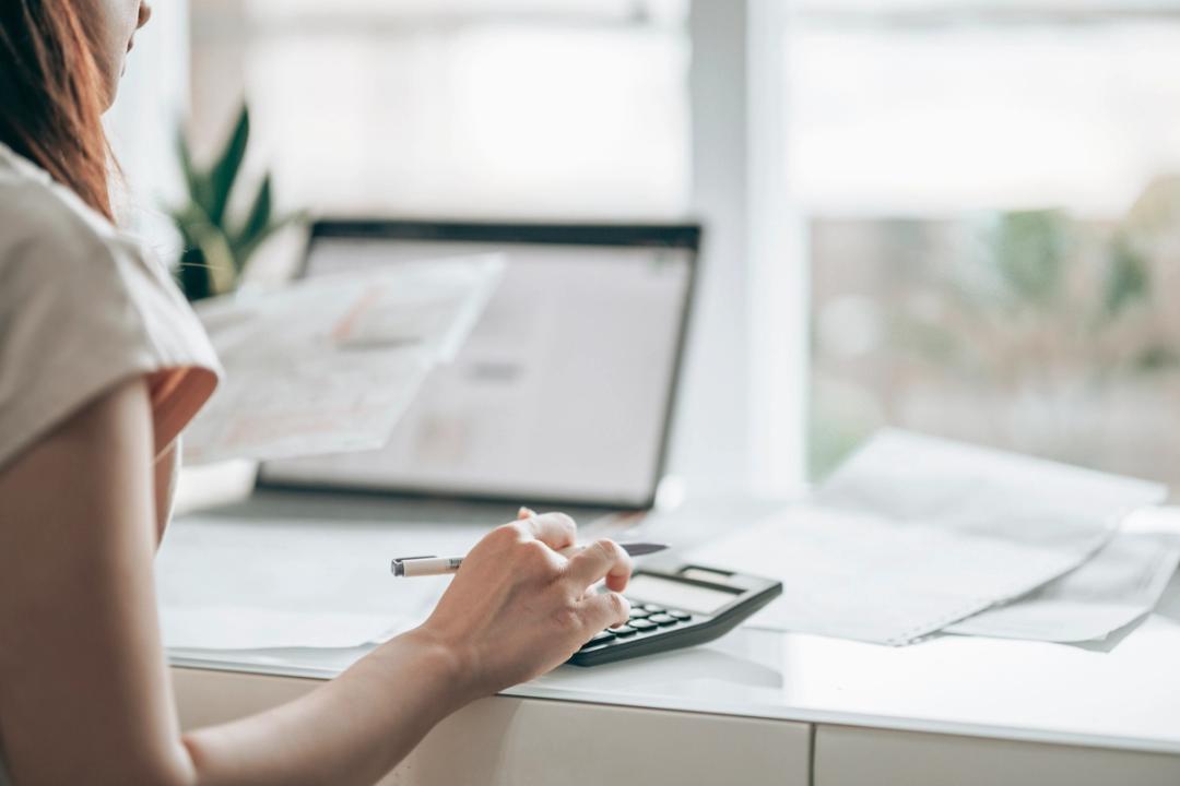 Woman using a calculator while looking at her laptop and paperwork