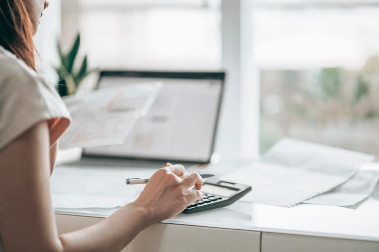 Woman using a calculator while looking at her laptop and paperwork