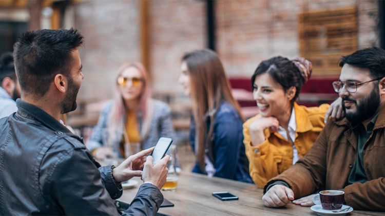 Group of young and adult men and women smiling in casual clothing, enjoying drinks around a table, sitting outdoors.