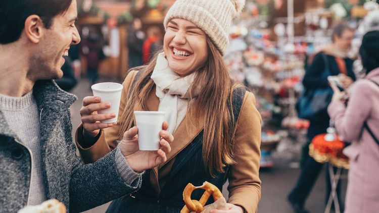 Smiling adult man and woman holding coffees and pretzel in an outdoor winter setting.