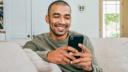Adult man smiling while casually dressed sat on sofa looking at smart phone.