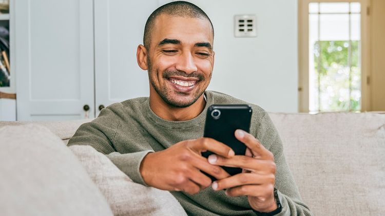Adult man smiling while casually dressed sat on sofa looking at smart phone.