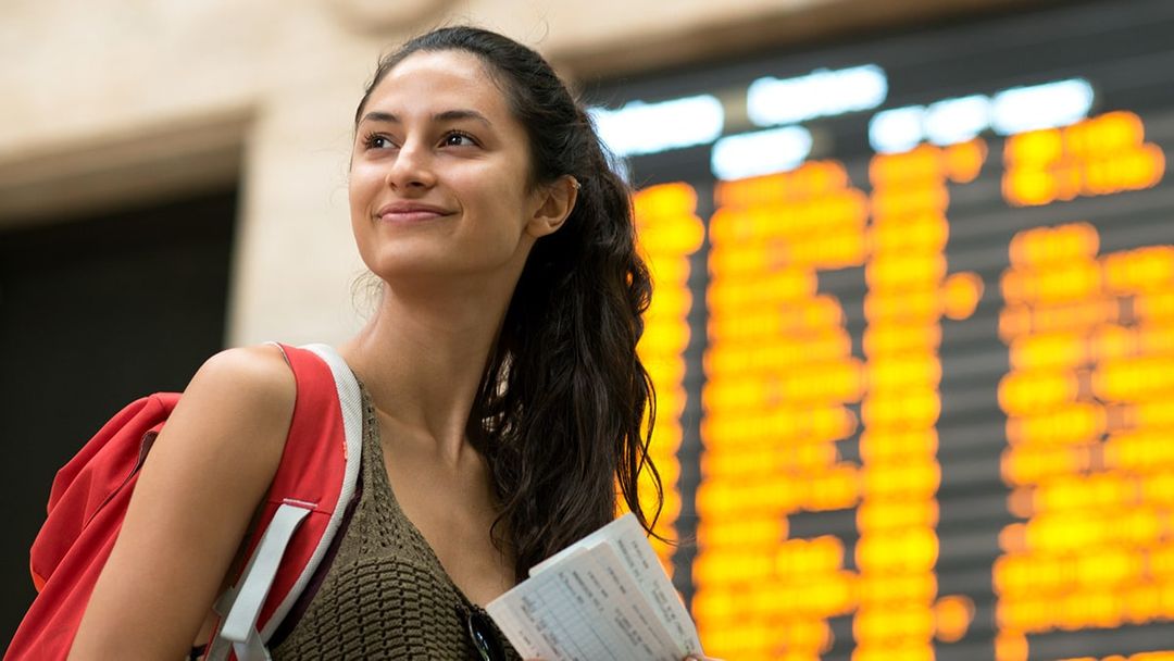 Young adult female smiling while on a travel journey, with airport departures board blurred in the background.
