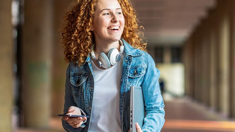 Cheerful young adult woman with headphones around neck holding laptop under arm, enjoying the outdoors lifestyle.