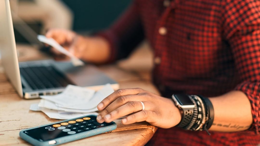 Close-up of one young adult man using calculator on mobile phone with human hand on table indoors.