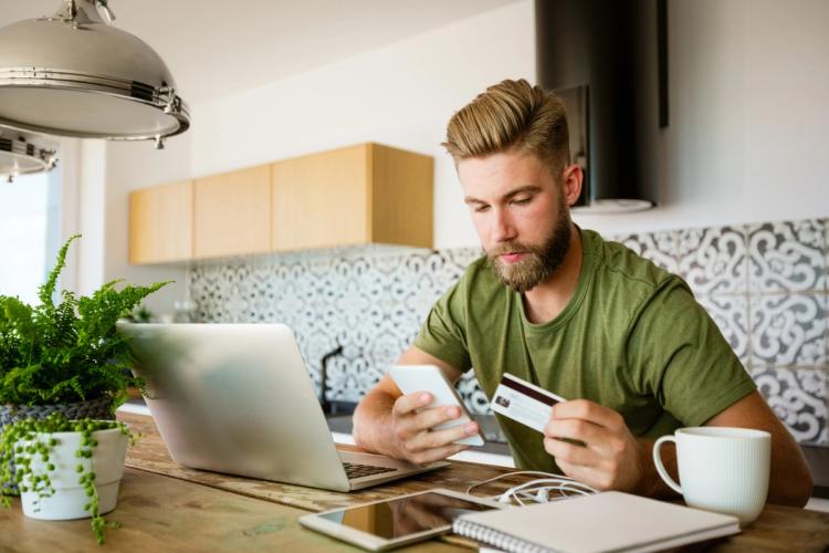 Man looking at his phone and credit card while sat in front of a laptop