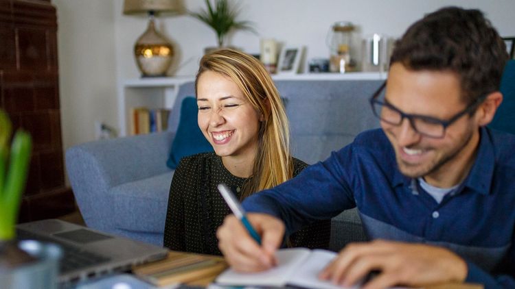 Indoors, smiling man and woman sat at coffee table, working together at laptop.
