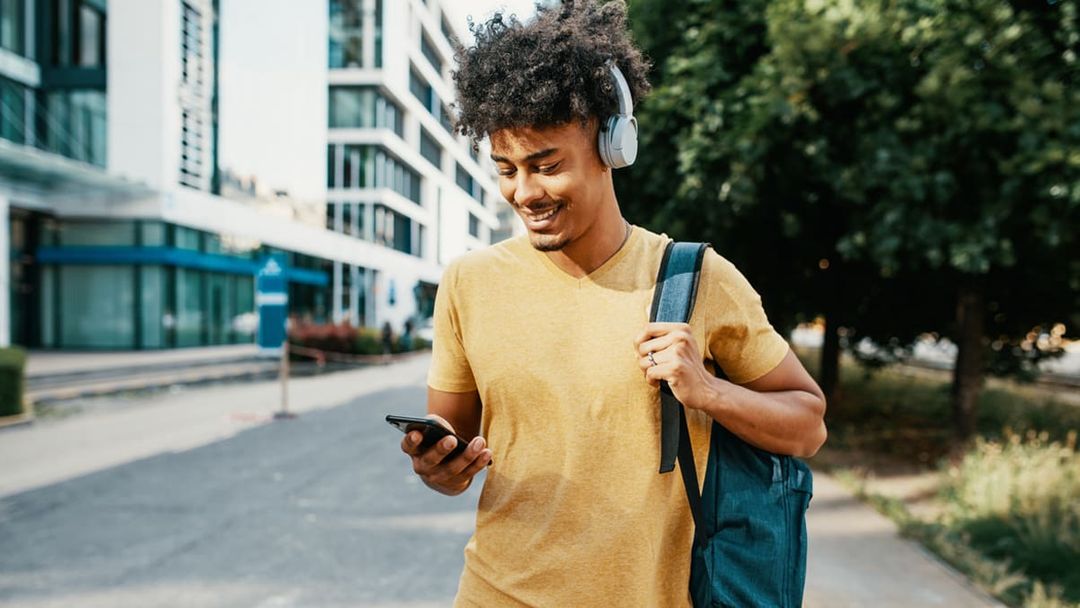 Young man wearing headphones walking with backpack and looking at mobile phone