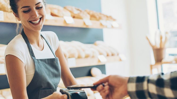 Adult female smiling in apron, working in a bakery and helping a customer at the till.