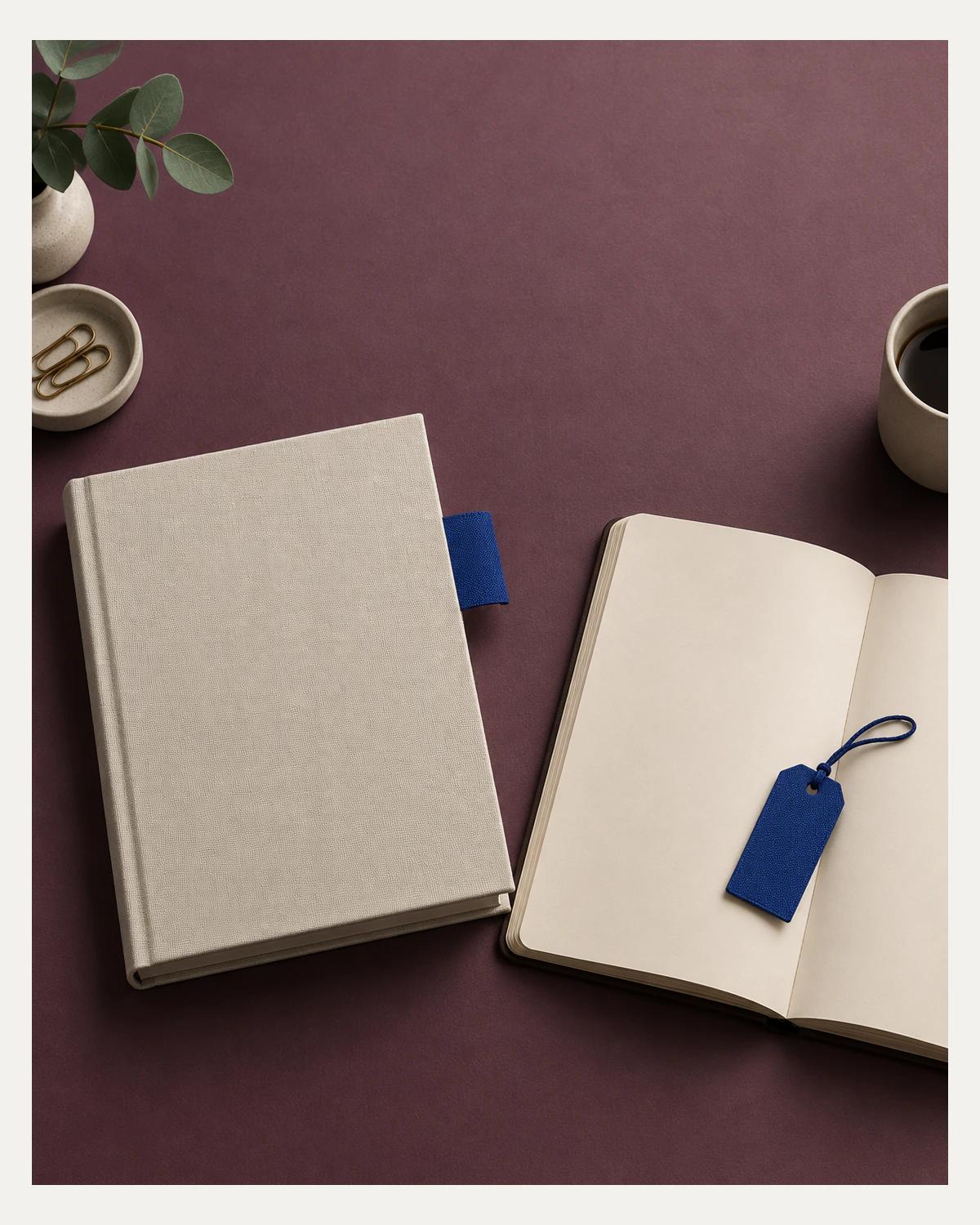 A closed cream book with a blue fabric tab and an open blank notebook with a loose blue tag sit on a plum-coloured desk.