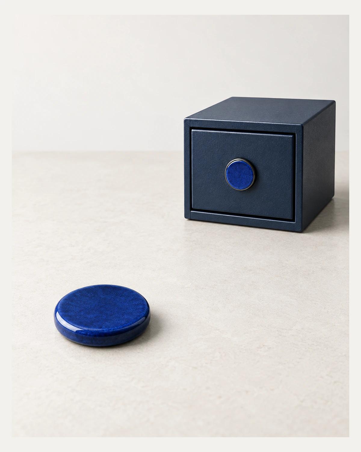 A cobalt ceramic token rests on a pale desk beside a navy metal drawer box with a matching blue circular inset.