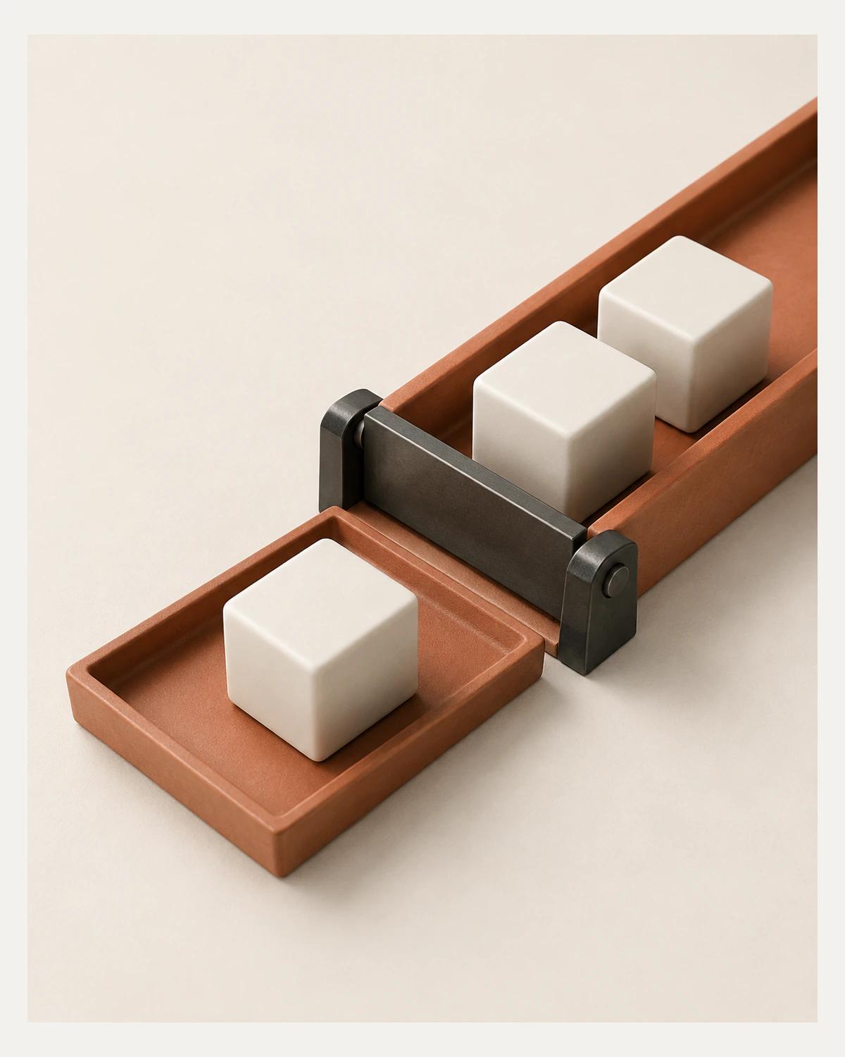 Two ivory blocks wait in a terracotta channel behind a lowered graphite gate while a third rests on a separate output tray.
