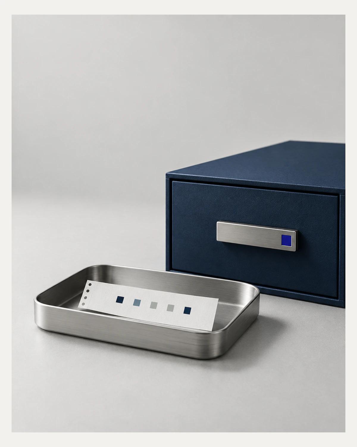 A small perforated paper record in an aluminium tray beside a closed navy document drawer.