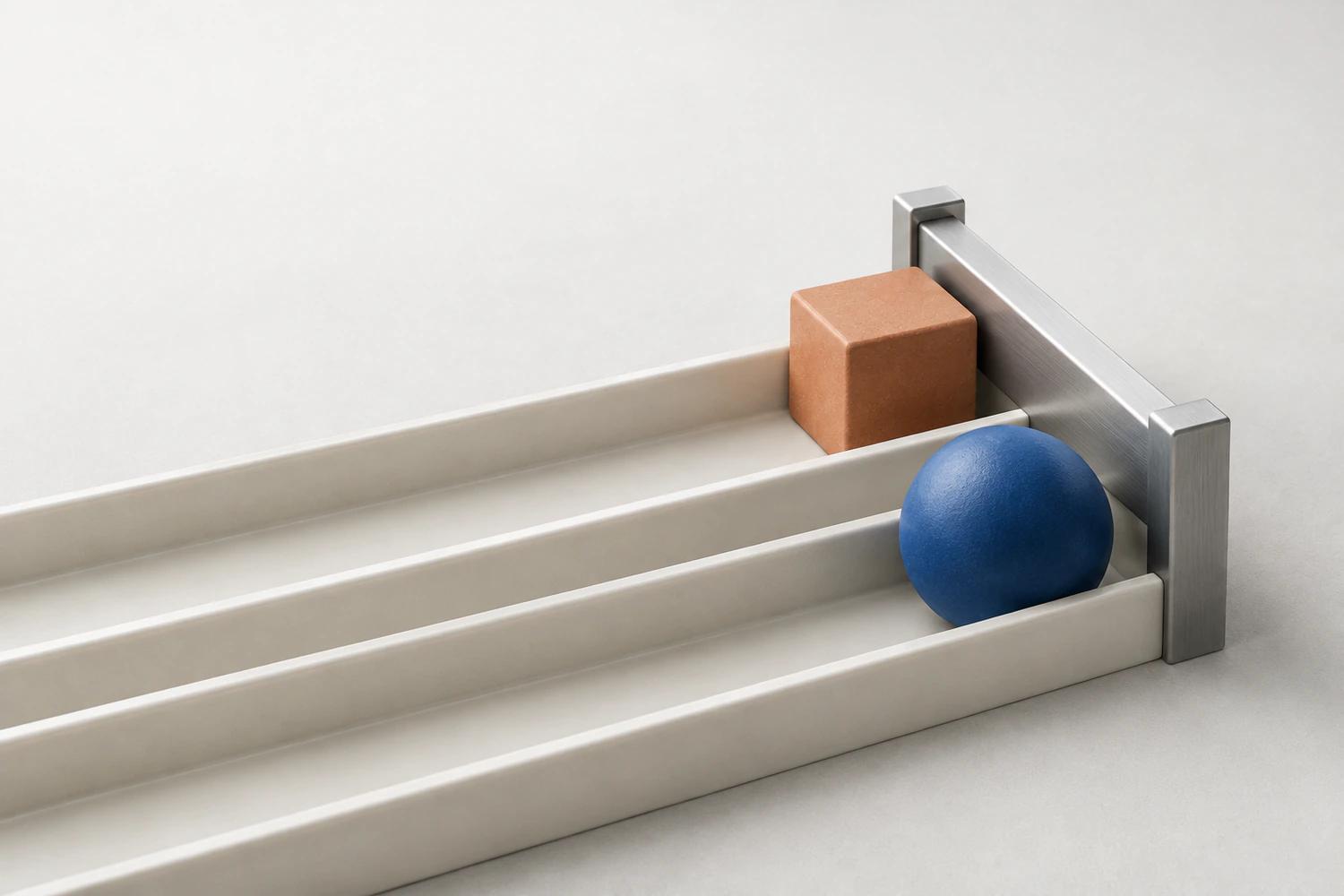 A cobalt sphere and a terracotta cube sit in two ivory channels against one lowered brushed-metal bar.