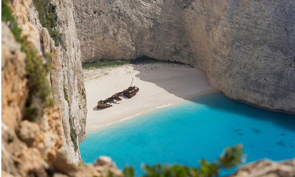 En strand i Hellas med et forlatt skip. Blått vann og høye fjell omkranser stranden.