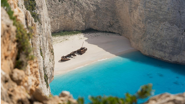 En strand i Hellas med et forlatt skip. Blått vann og høye fjell omkranser stranden.