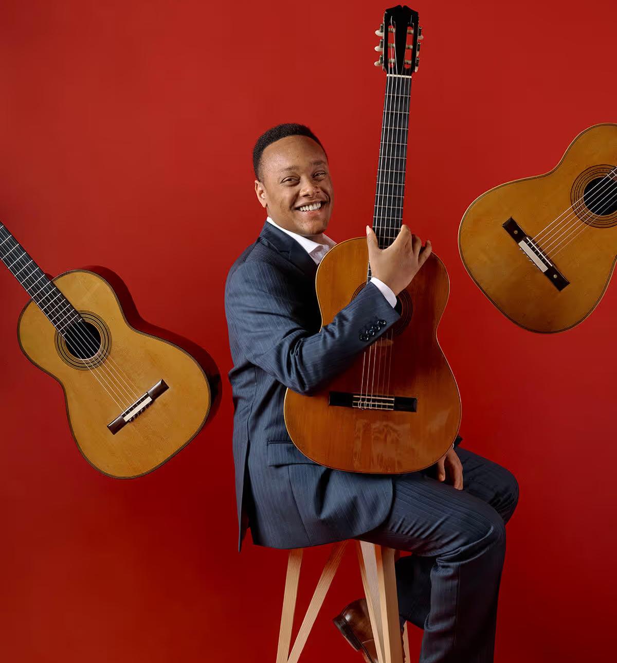 Raphael with his guitar in front of a red background with guitars floating around him.