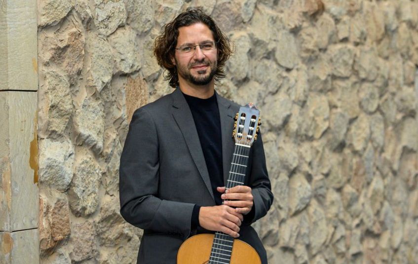 Jose Maria in front of a stone wall in a fancy suit holding his guitar.