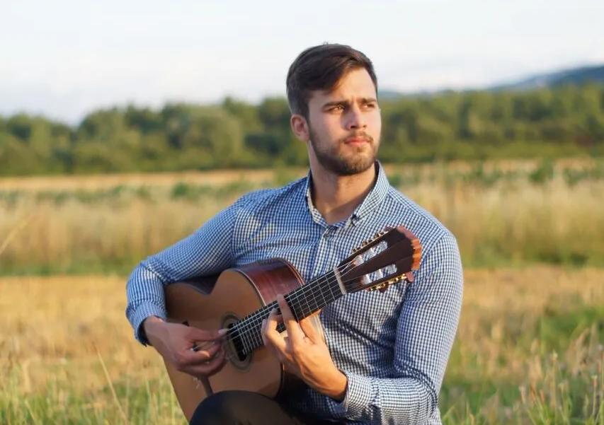 Ivan sitting on a round by a field playing the guitar.
