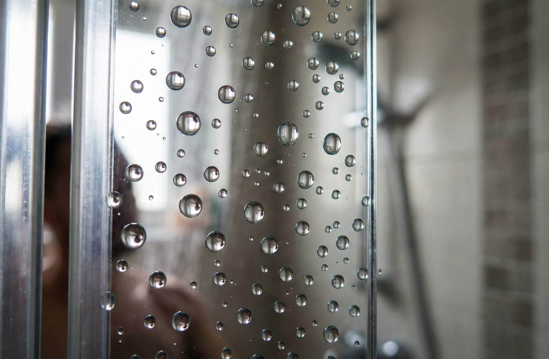 Primo piano di gocce d'acqua perfettamente sferiche che scivolano via da un vetro doccia trattato con nanotecnologia ECTOR B-10, senza lasciare traccia.