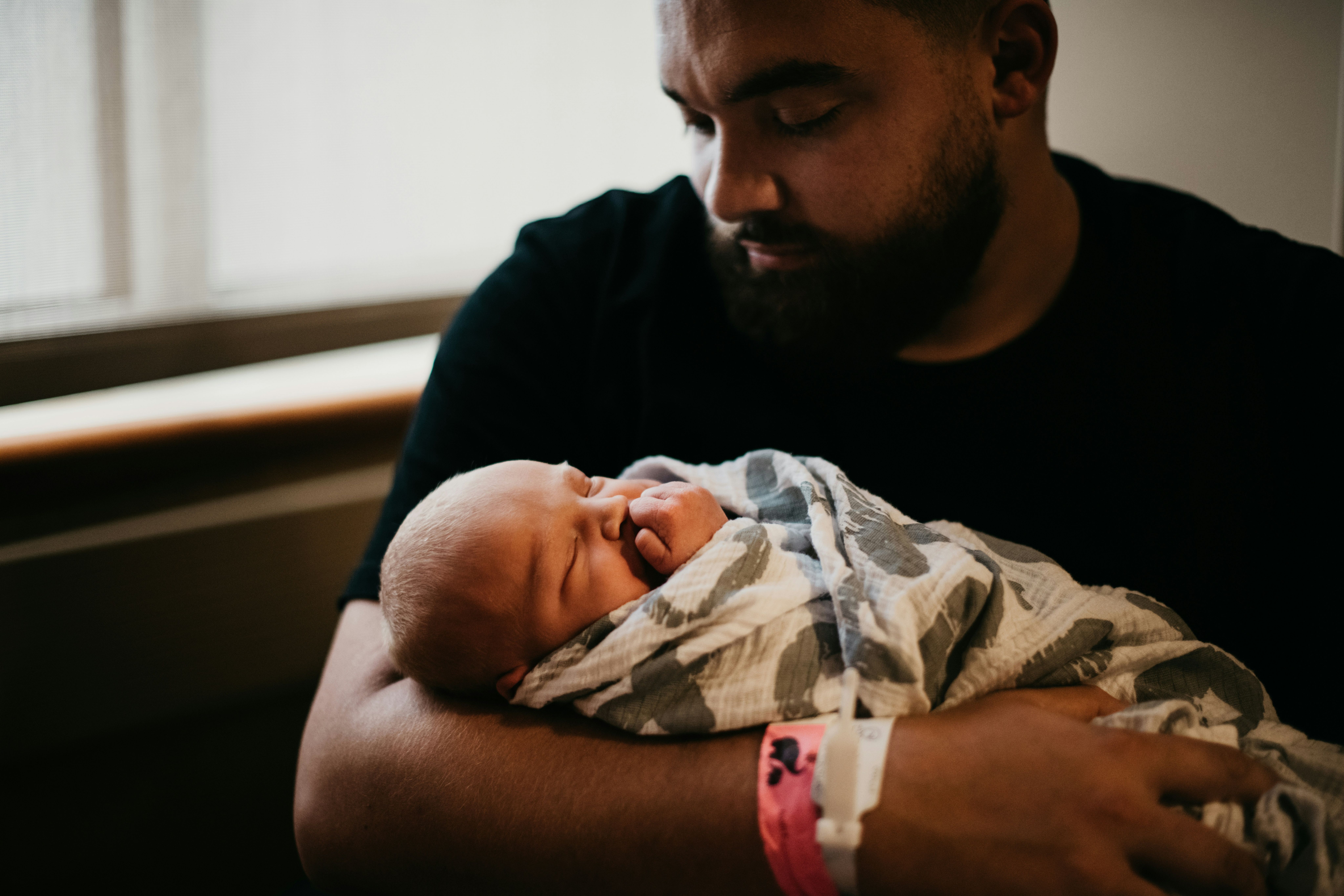 Father in hospital wristbands gazing down at his swaddled newborn, reflecting a dad coping after his baby's birth injury.