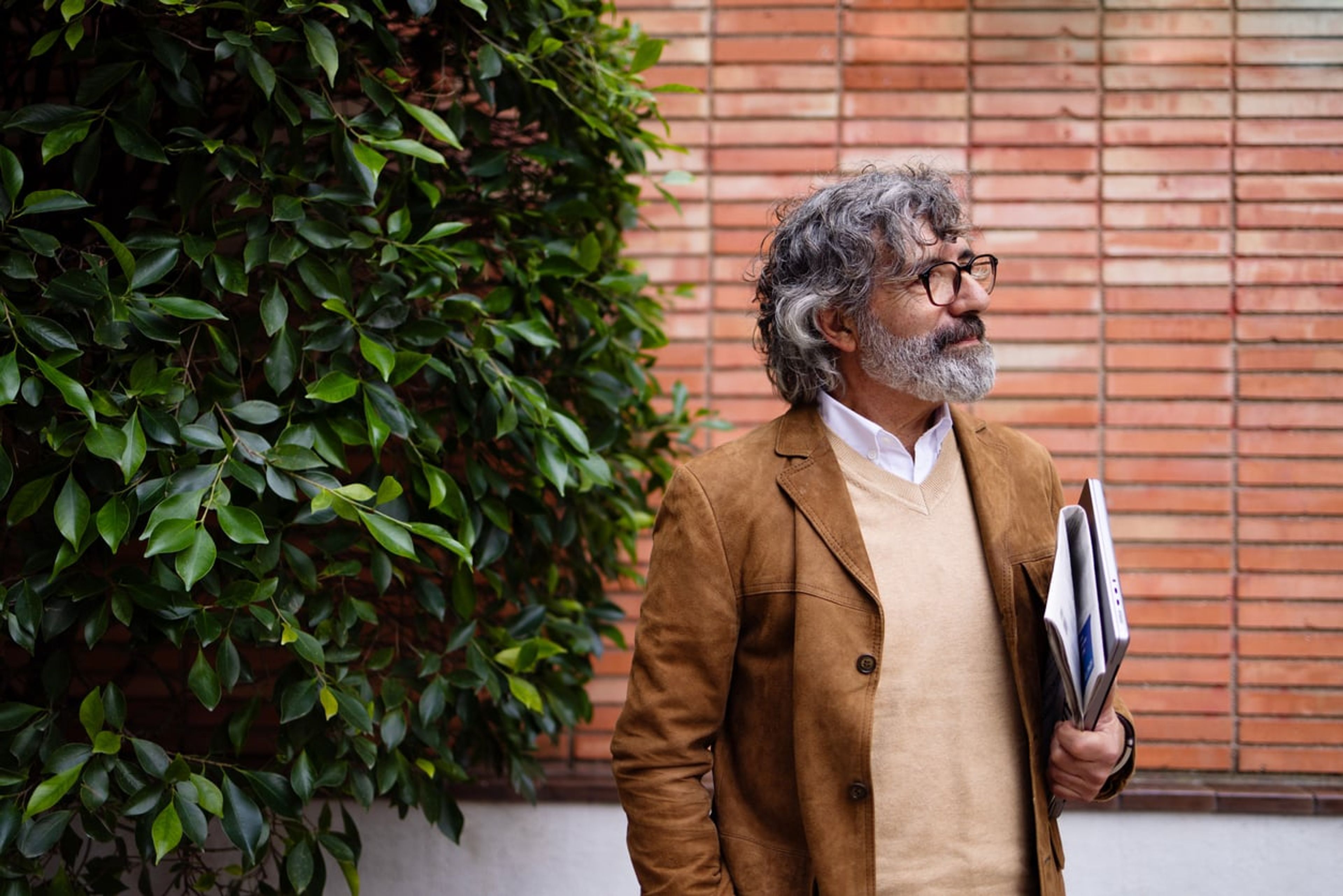 A mature white professor stands outdoors by a brick wall and a tree holding a laptop and documents.