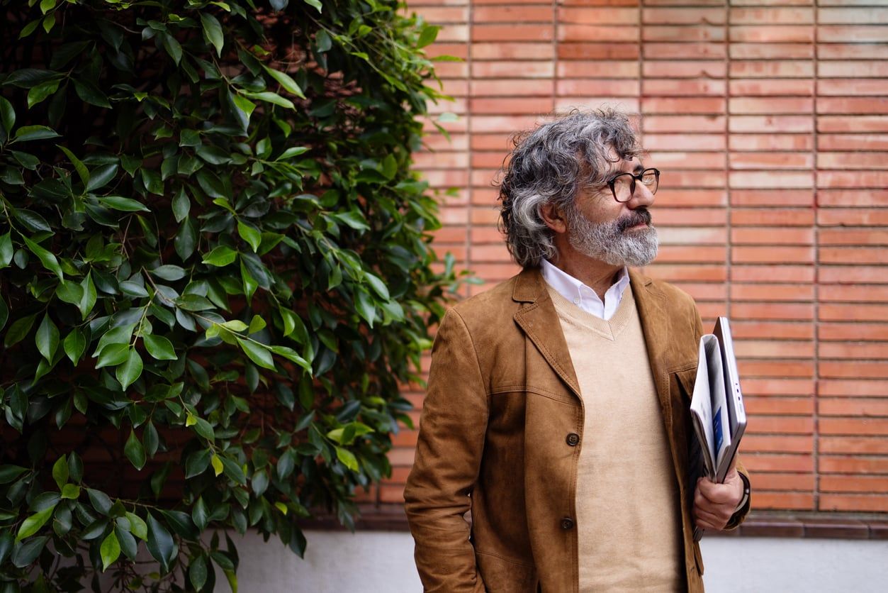 A mature white professor stands outdoors by a brick wall and a tree holding a laptop and documents.