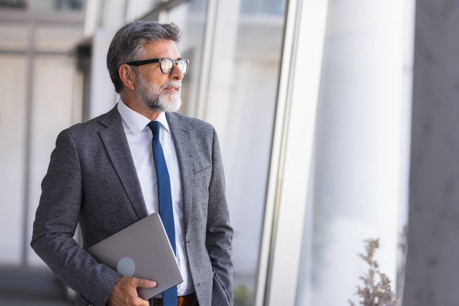 A senior executive holding a laptop and looking out an office window.