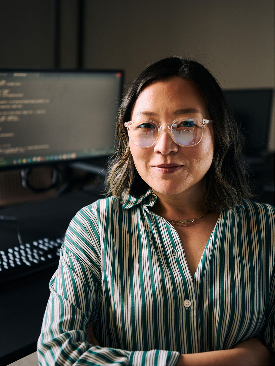 An engineer standing with arms crossed at a desk with code on screen