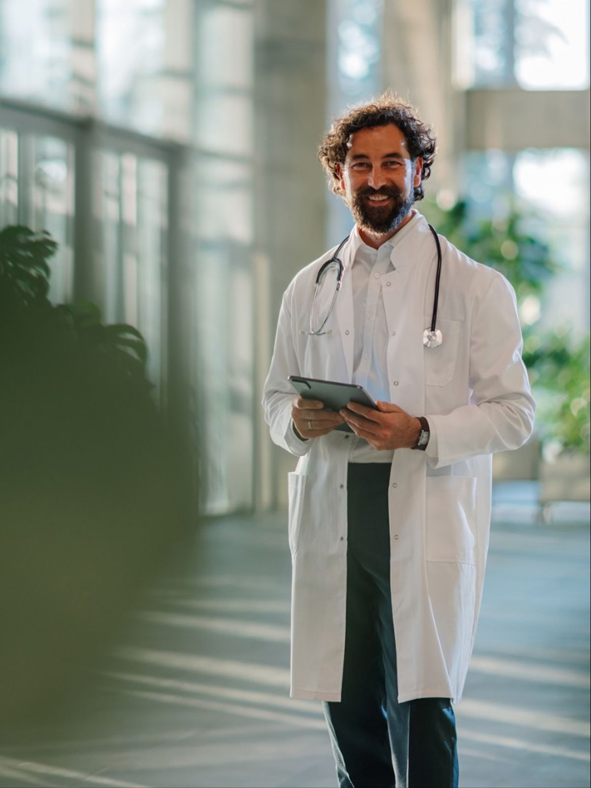 A smiling physician holding a tablet in a hospital corridor