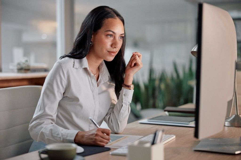 A professional working at a desk, reviewing documents on a computer screen in a bright office.