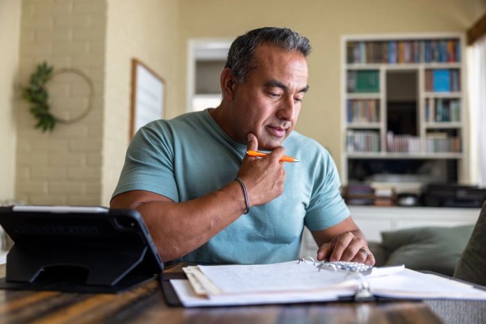 A man sits at a desk looking at a binder of papers as he uses a calculator.