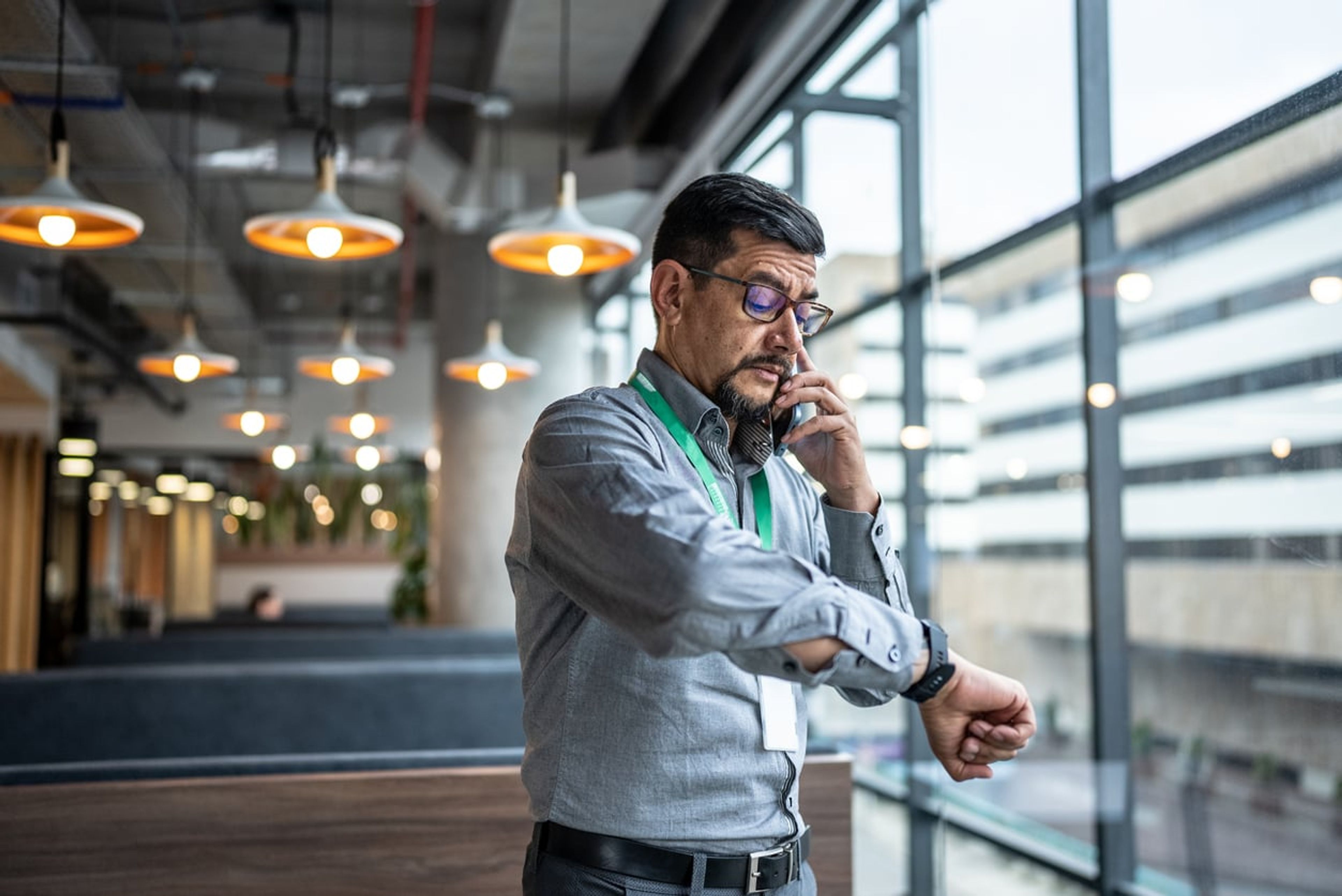 A mature man in a blue shirt is talking on the phone while checking the time on his watch.