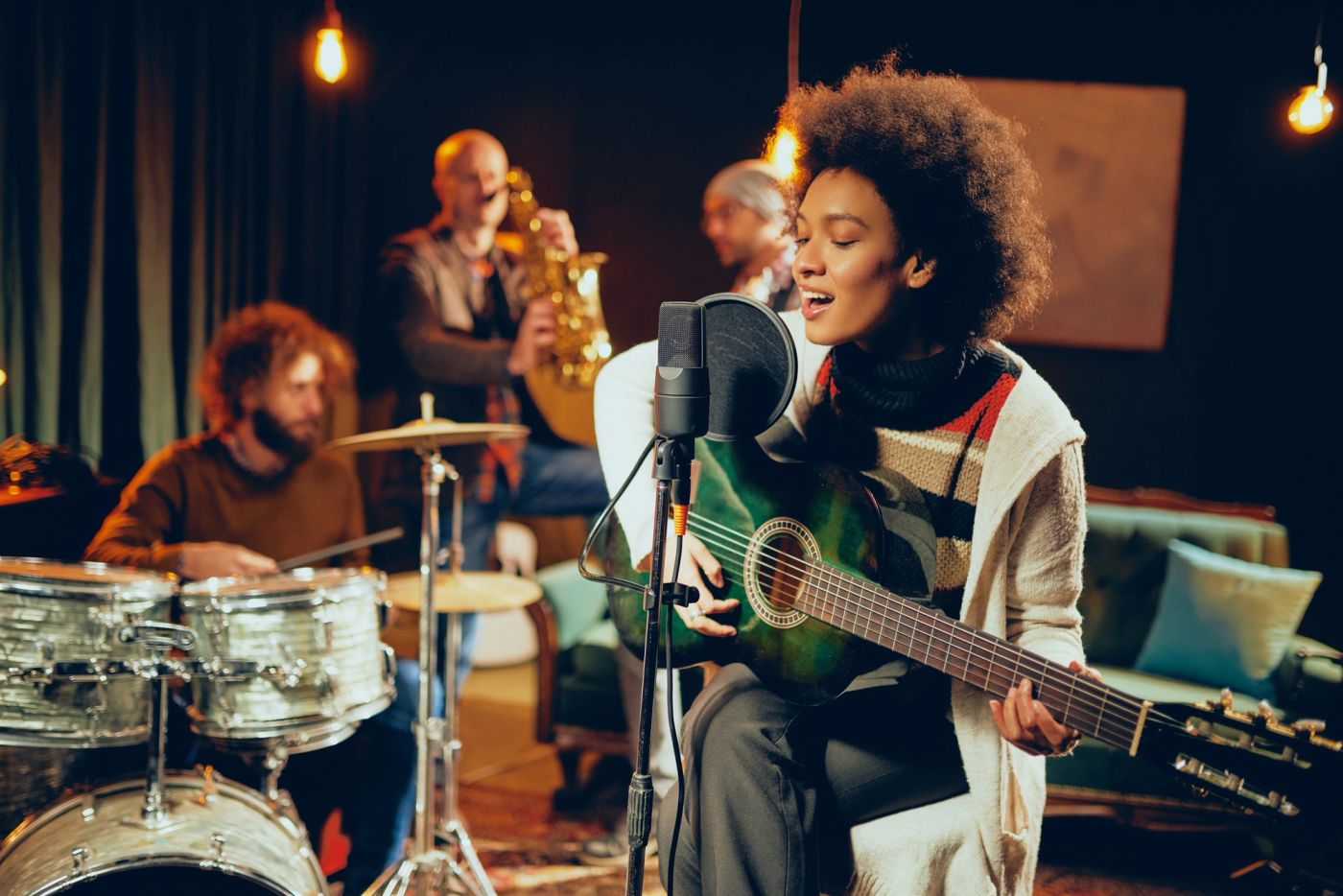 woman singing and playing guitar while sitting on chair with legs crossed. In background drummer, saxophonist and bass guitarist.