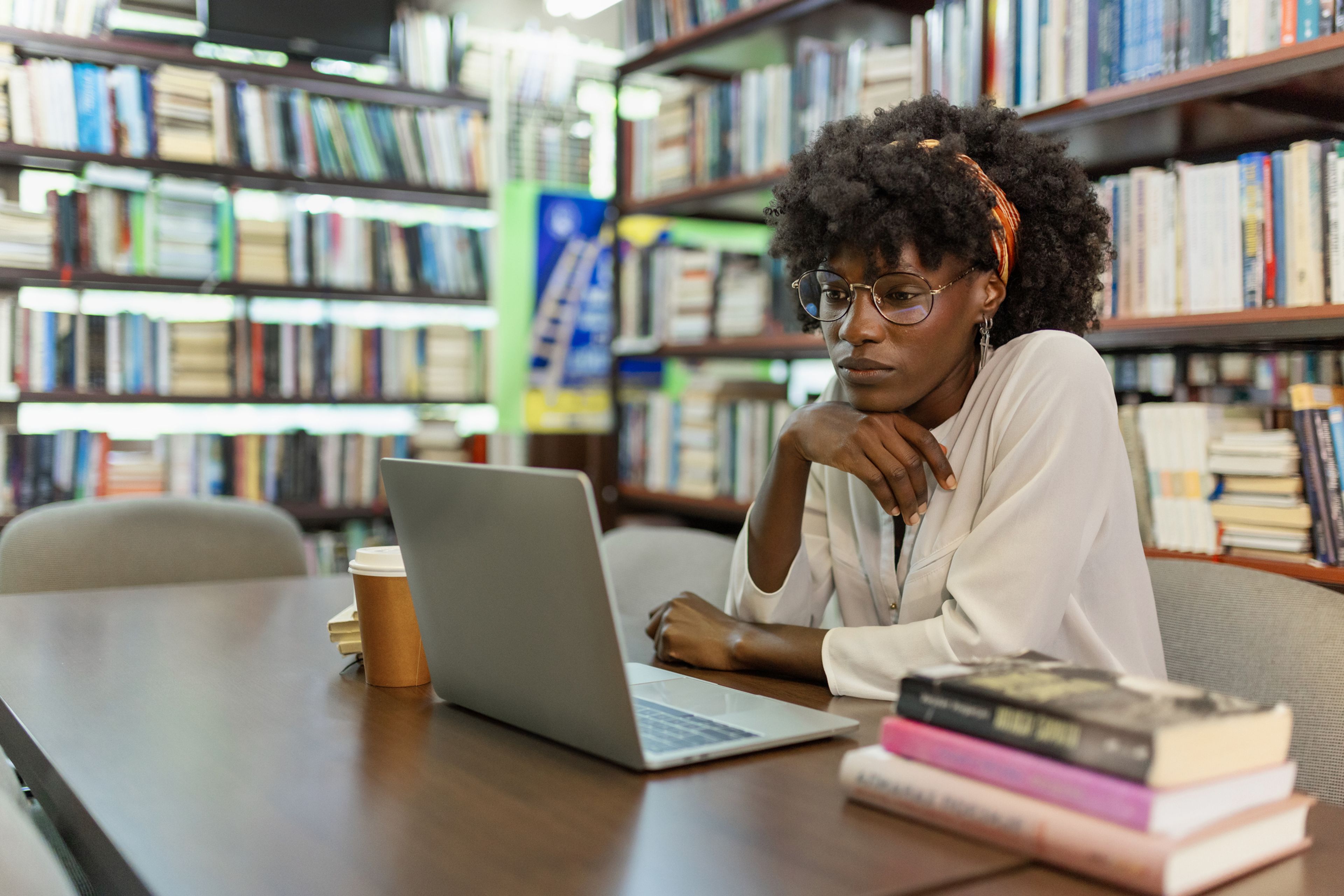 A researcher working on a laptop at a table surrounded by books in a library.